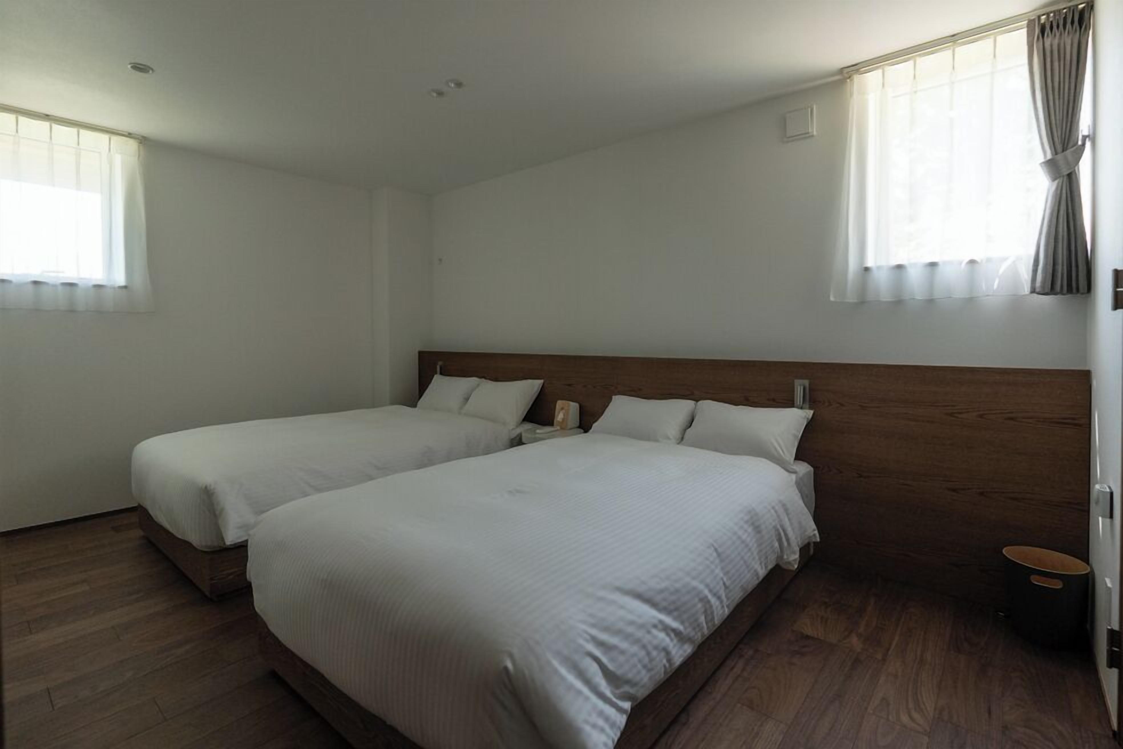 Een hotelkamer met twee enkele bedden, een houten hoofdbord, witte beddengoed, een klein nachtkastje en een raam met gordijnen.