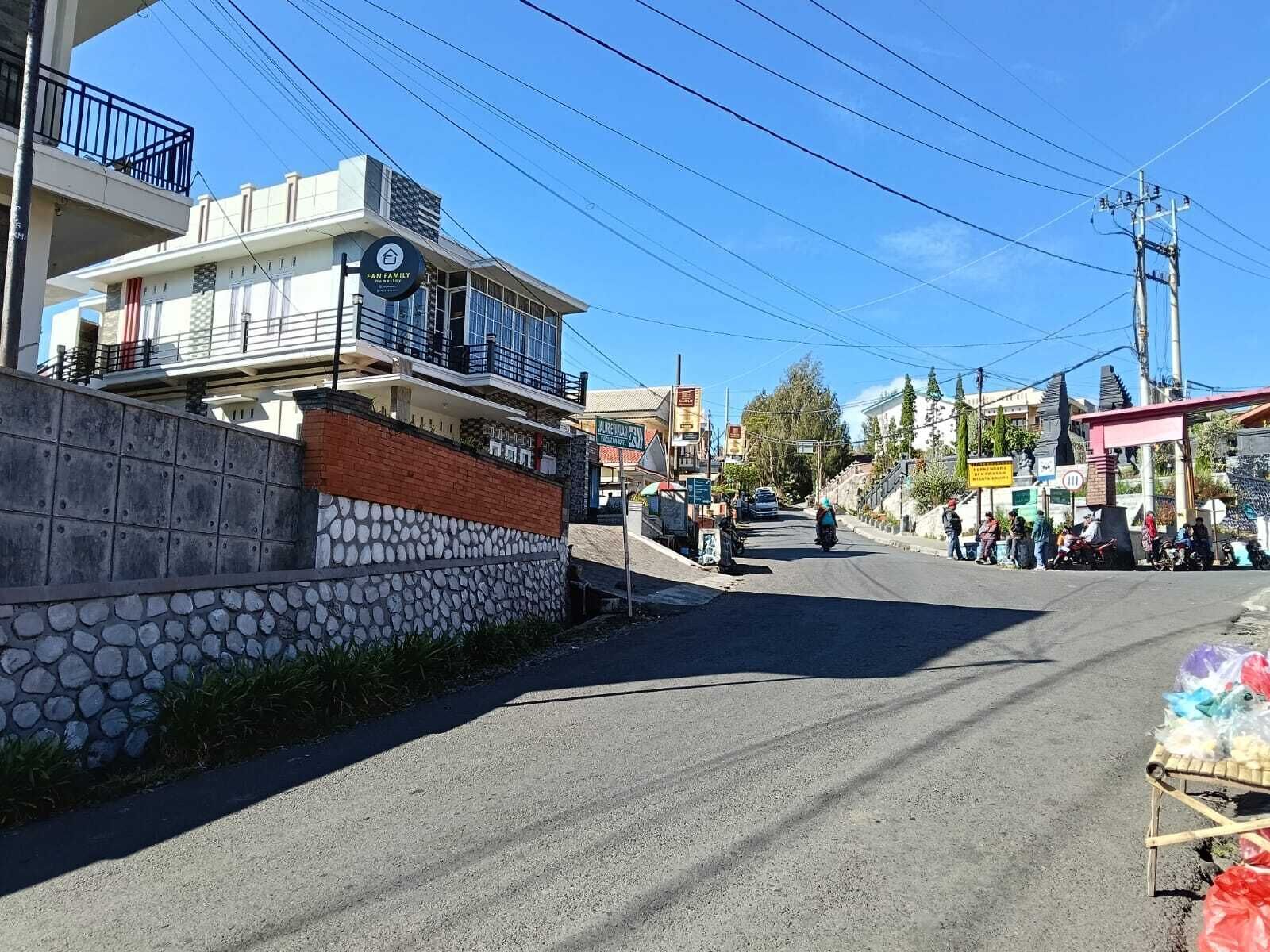 Una vista di strada con un edificio con la scritta 'Fan Family' a sinistra, un muro di pietra e un piccolo negozio a destra.