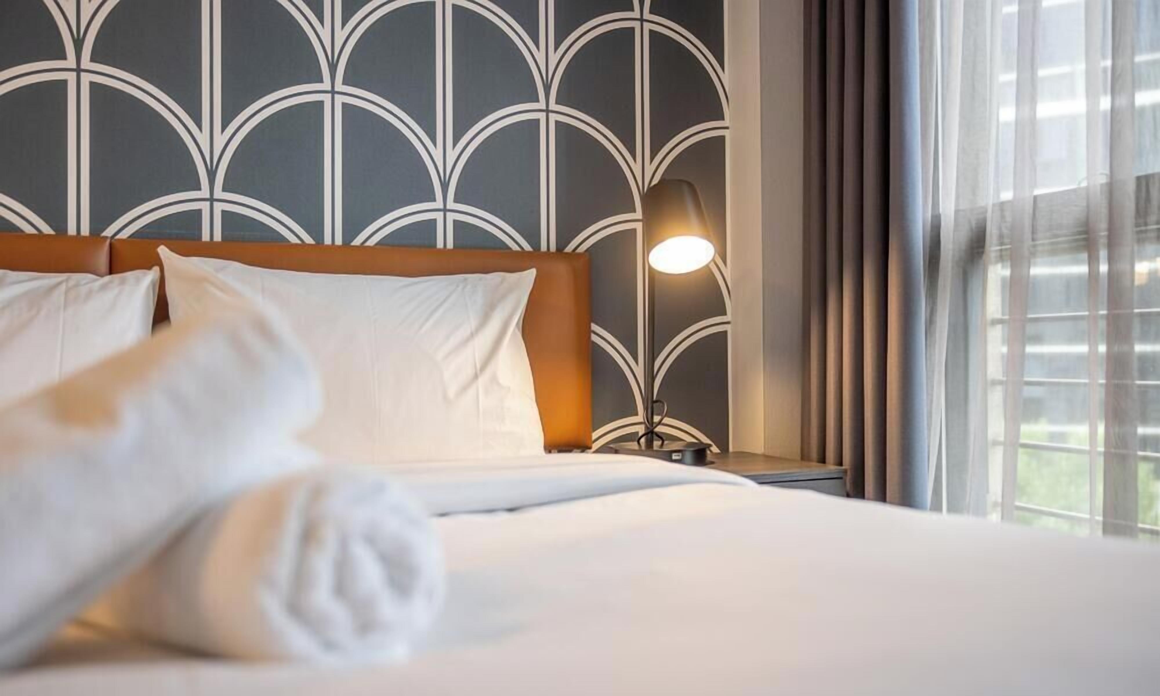 Habitación de hotel con cama, almohadas, mesita de noche con lámpara y ventana con cortinas.