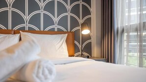 Habitación de hotel con cama, almohadas, mesita de noche con lámpara y ventana con cortinas.