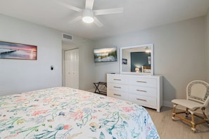 Une chambre à coucher comprenant un lit, une commode, une chaise, un ventilateur de plafond et deux téléviseurs fixés au mur.