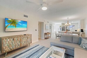A modern living room with a large flat-screen TV, a sofa, a dining area, and a kitchen in the background. - Gulf Breezes at Runaway Bay 147 (Bradenton Beach)