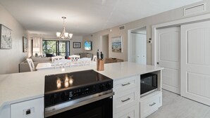Eine moderne Küche mit Kochinsel, integriertem Backofen und Mikrowelle. Es gibt einen Essbereich mit weißem Tisch und Stühlen sowie ein Wohnzimmer mit Sofa und Fernseher.