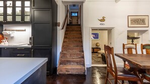 Eine Küche mit dunklen Schränken, einer Holztreppe, einem Essbereich mit Tisch und Stühlen sowie einem Flur, der zu einem Zimmer mit Fenster und einem gerahmten Bild an der Wand führt.