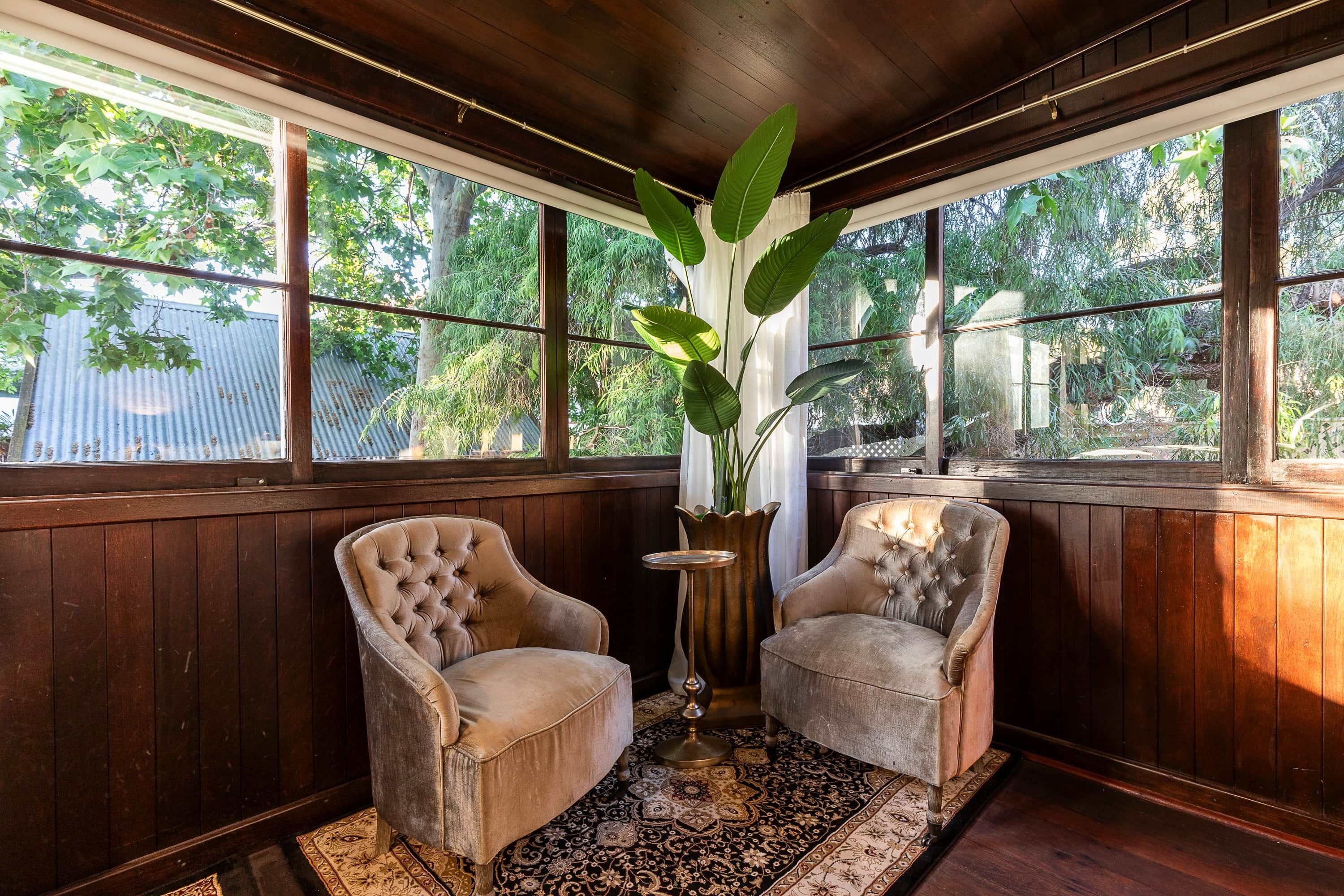 A room with two upholstered armchairs, a patterned rug, a potted plant, and large windows with a view of trees.