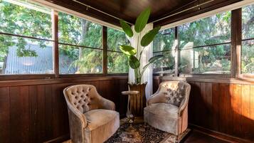 A room with two upholstered armchairs, a patterned rug, a potted plant, and large windows with a view of trees.