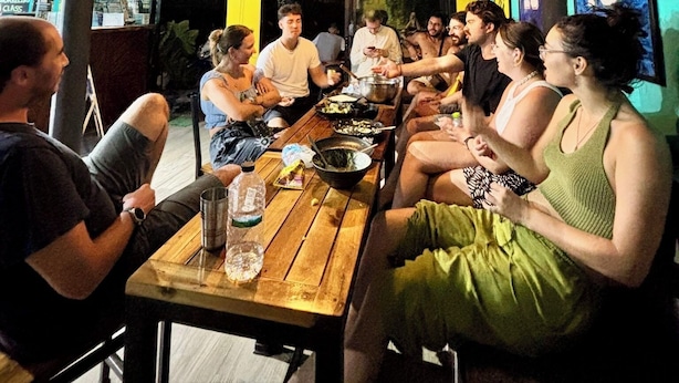 Μια ομάδα ανθρώπων γευματίζει σε ένα ξύλινο τραπέζι σε ένα δωμάτιο με τοίχους σε τιρκουάζ χρώμα και κρεμαστά φωτιστικά.