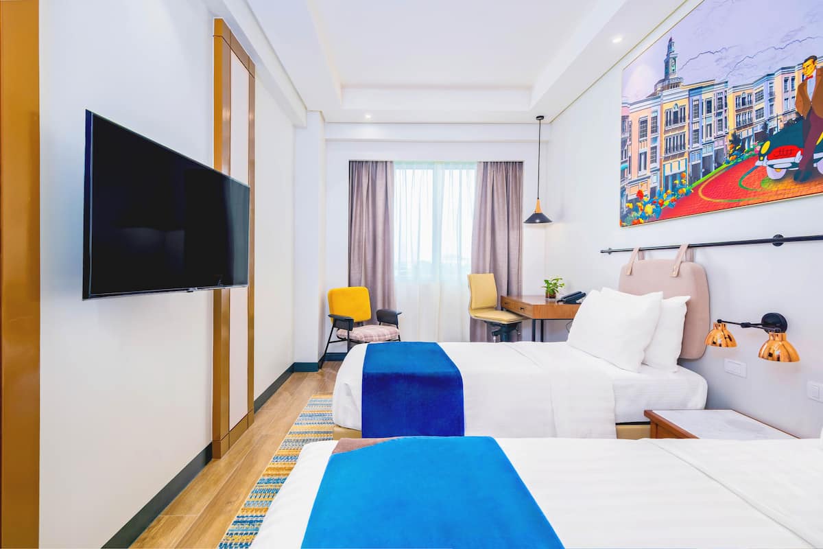 Una camera d'albergo con due letti, una televisione, una scrivania, una sedia, una lampada e un quadro appeso al muro.
