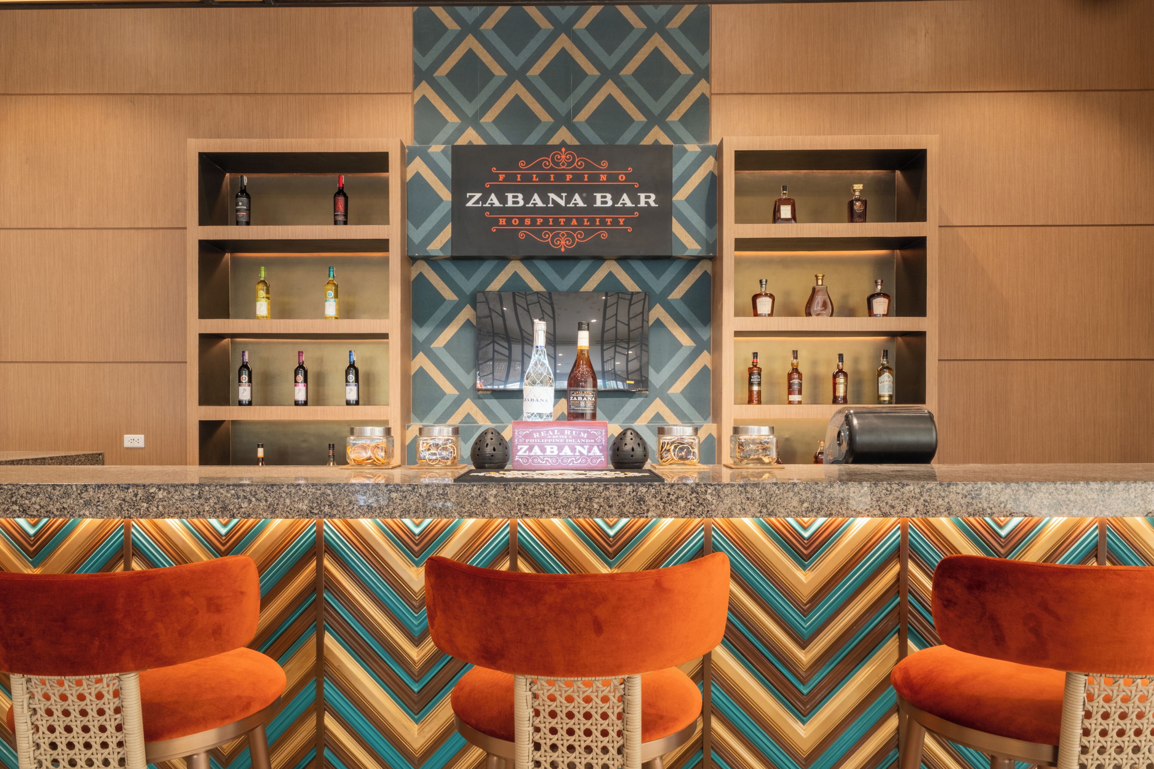A bar area with shelves of liquor bottles, a sign reading 'ZABANA BAR,' and three orange chairs.