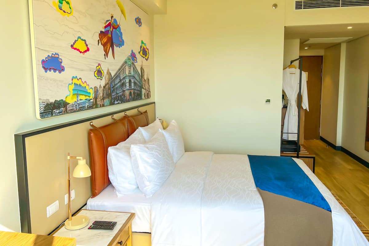 Una camera d'albergo con un letto, un comodino, una lampada, un quadro e un accappatoio appeso alla porta.