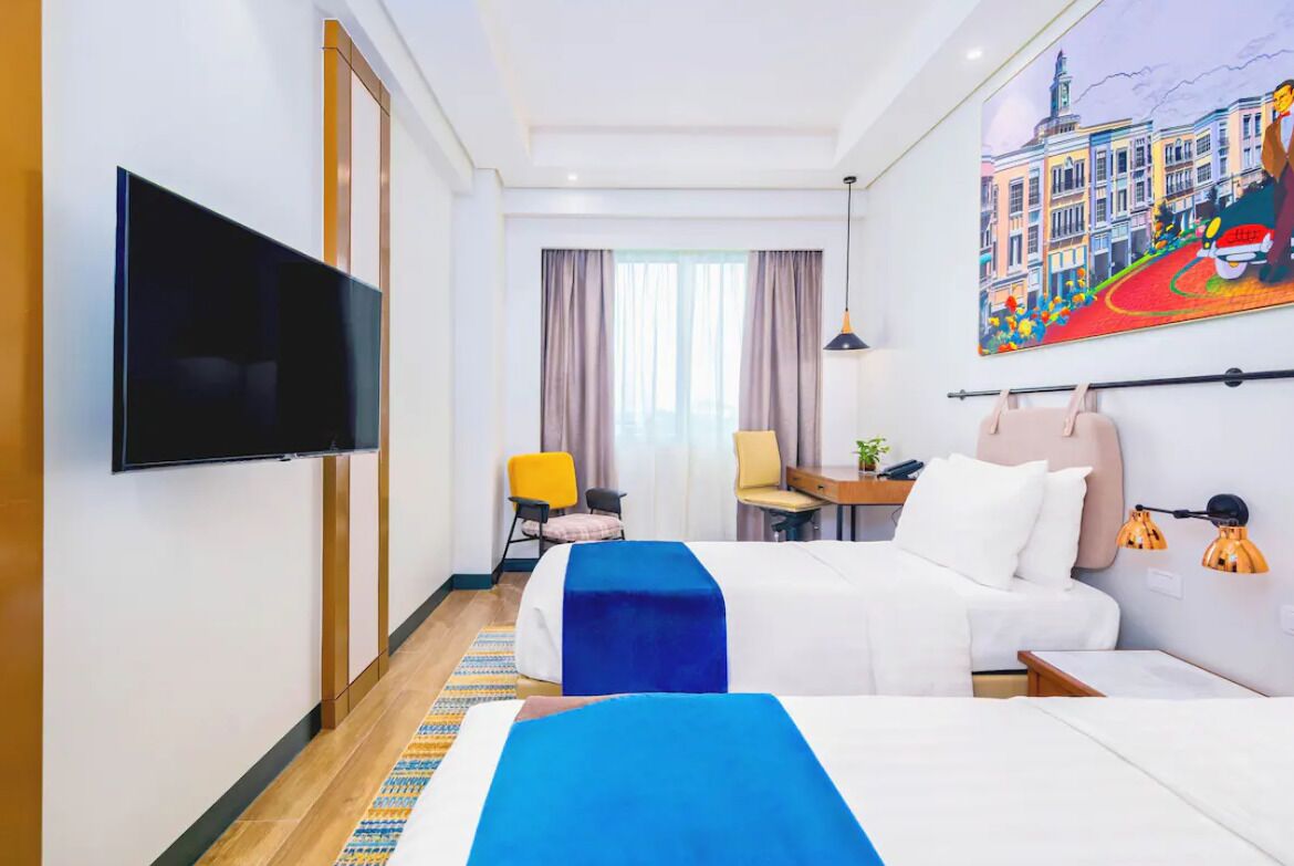 A hotel room with a TV, two beds, a desk, a chair, and a painting on the wall.