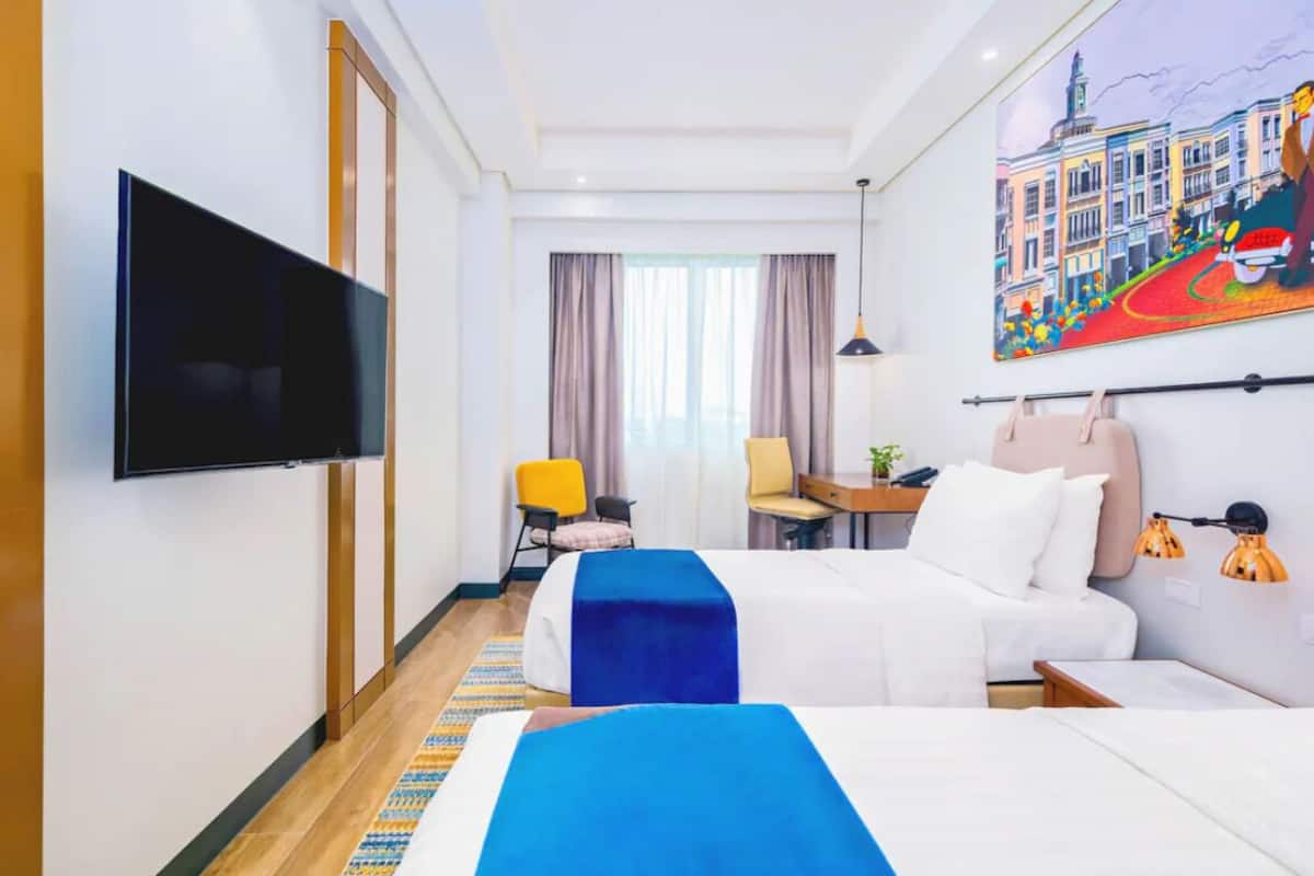 Una camera d'albergo con televisione, due letti, una scrivania, una sedia e un quadro appeso al muro.