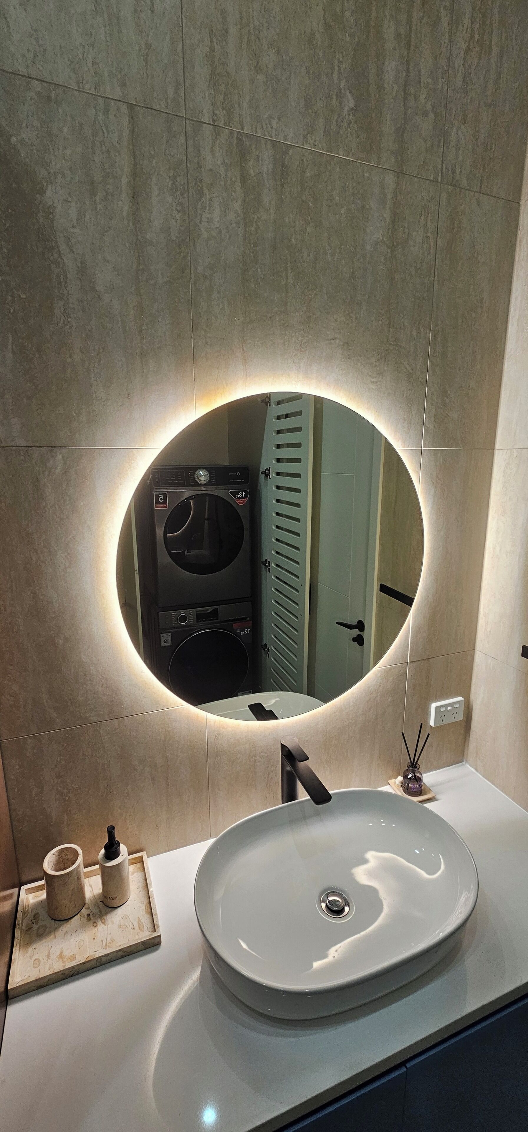 A bathroom with a round mirror, a sink, and a washing machine visible in the reflection.