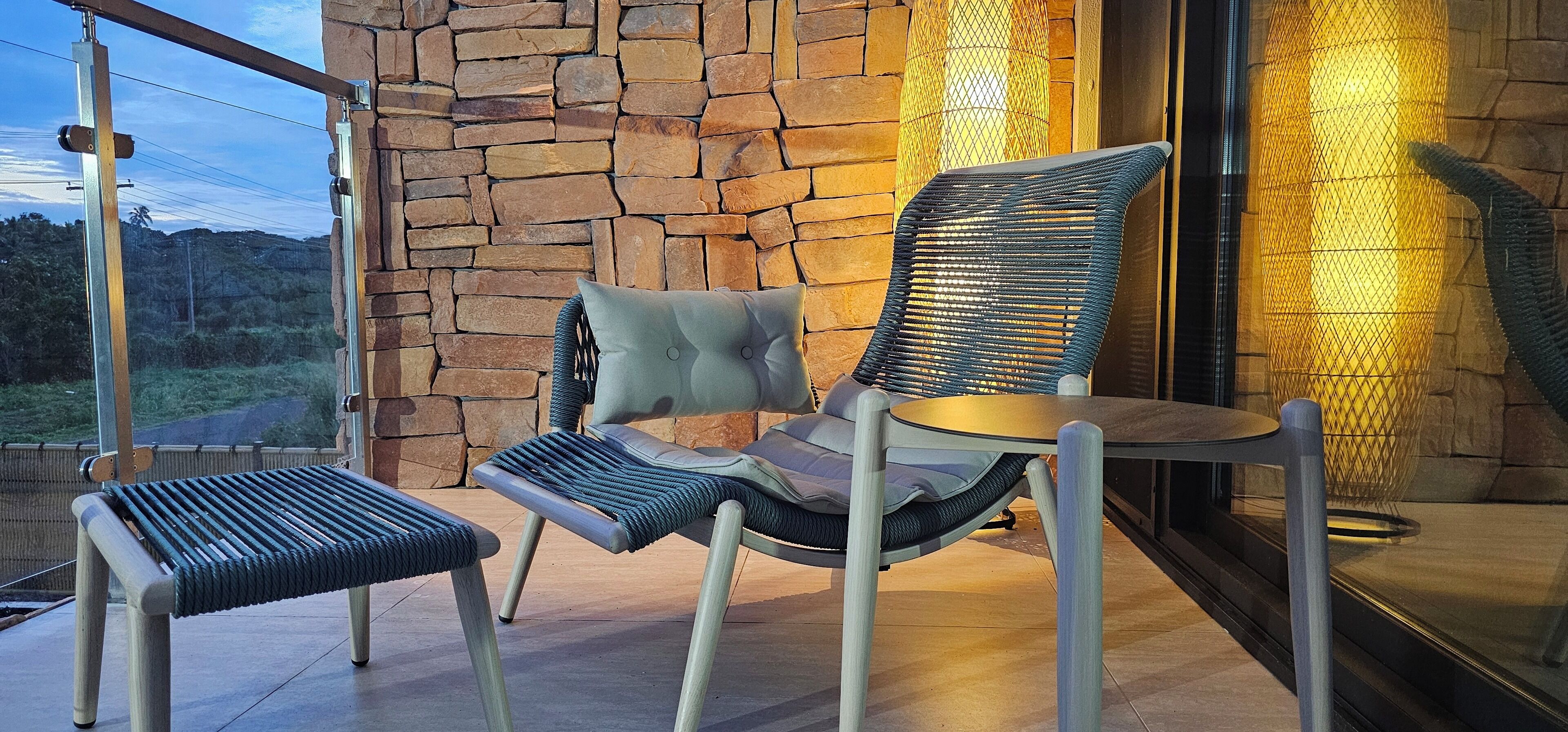 A modern outdoor seating area with wicker chairs and a small table, set against a stone wall and a glass door leading to a scenic view.