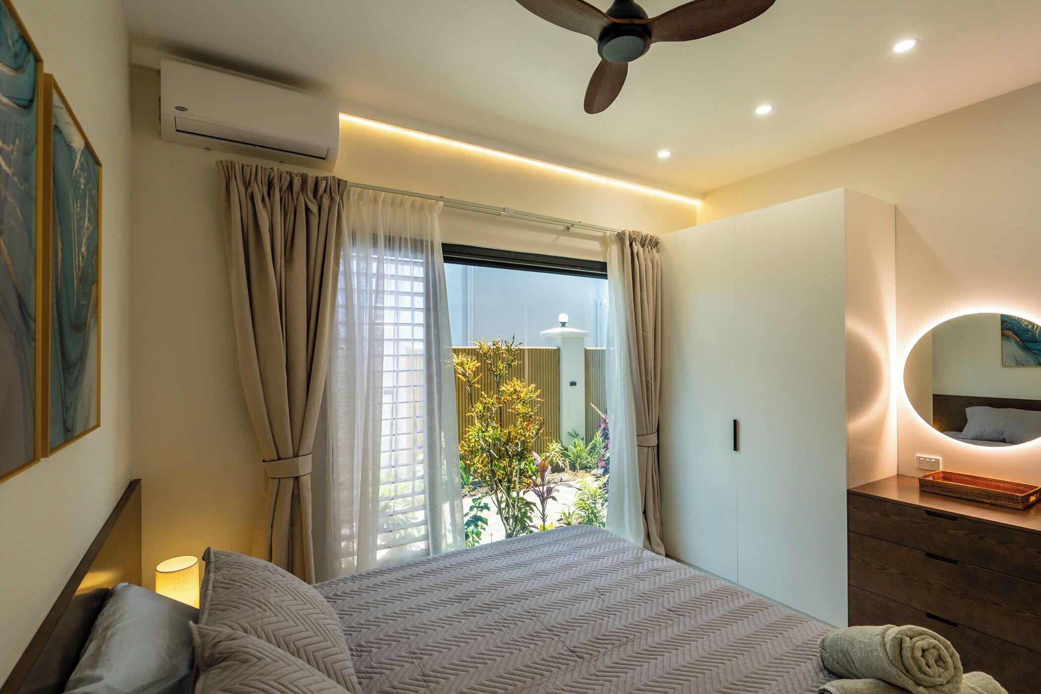A modern bedroom with a large bed, a ceiling fan, an air conditioning unit, a bedside lamp, a wooden dresser, a round mirror, and a view of greenery outside.