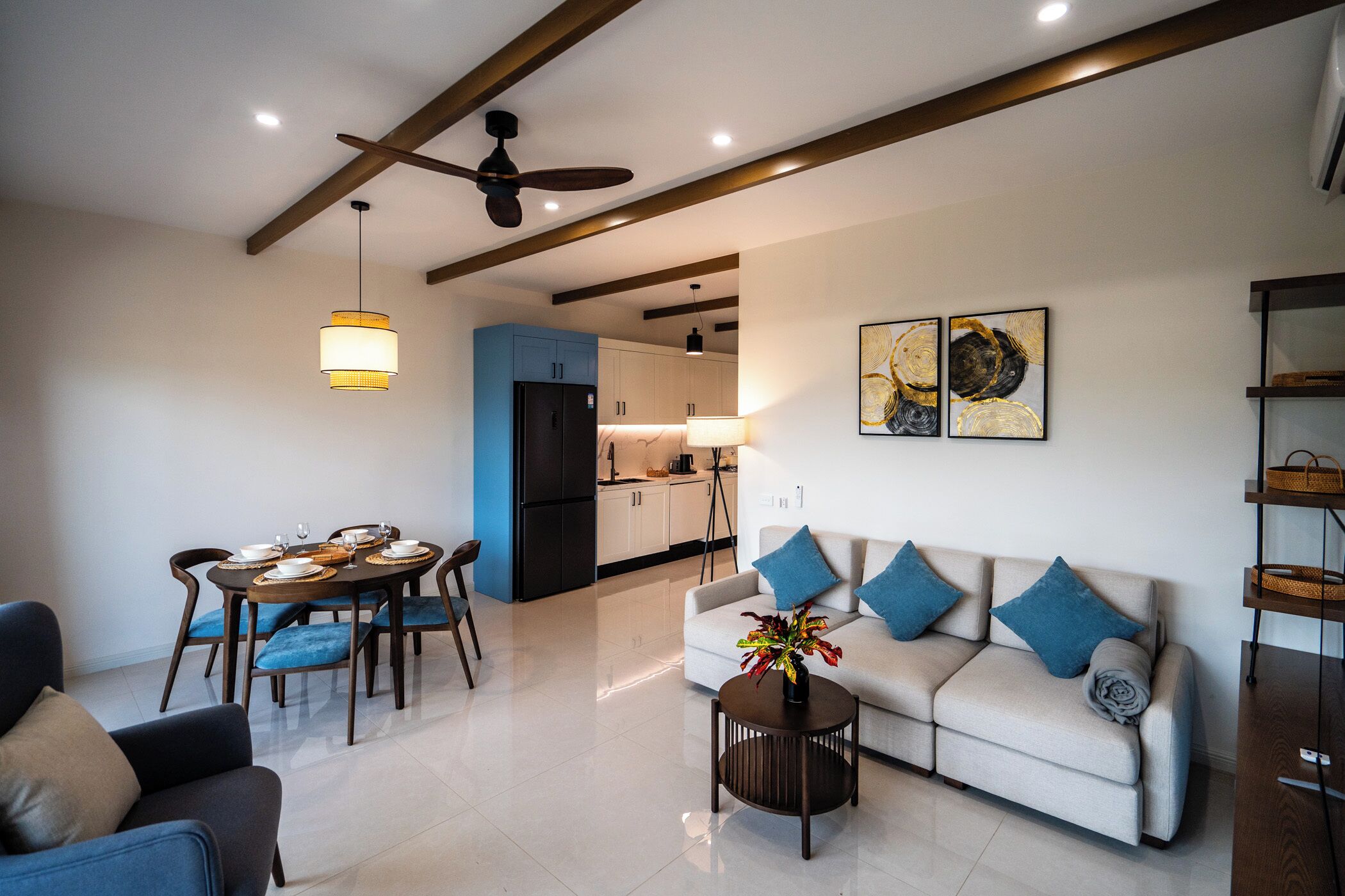 A modern living room with a sofa, dining table, and chairs, a kitchen in the background, and a ceiling fan.