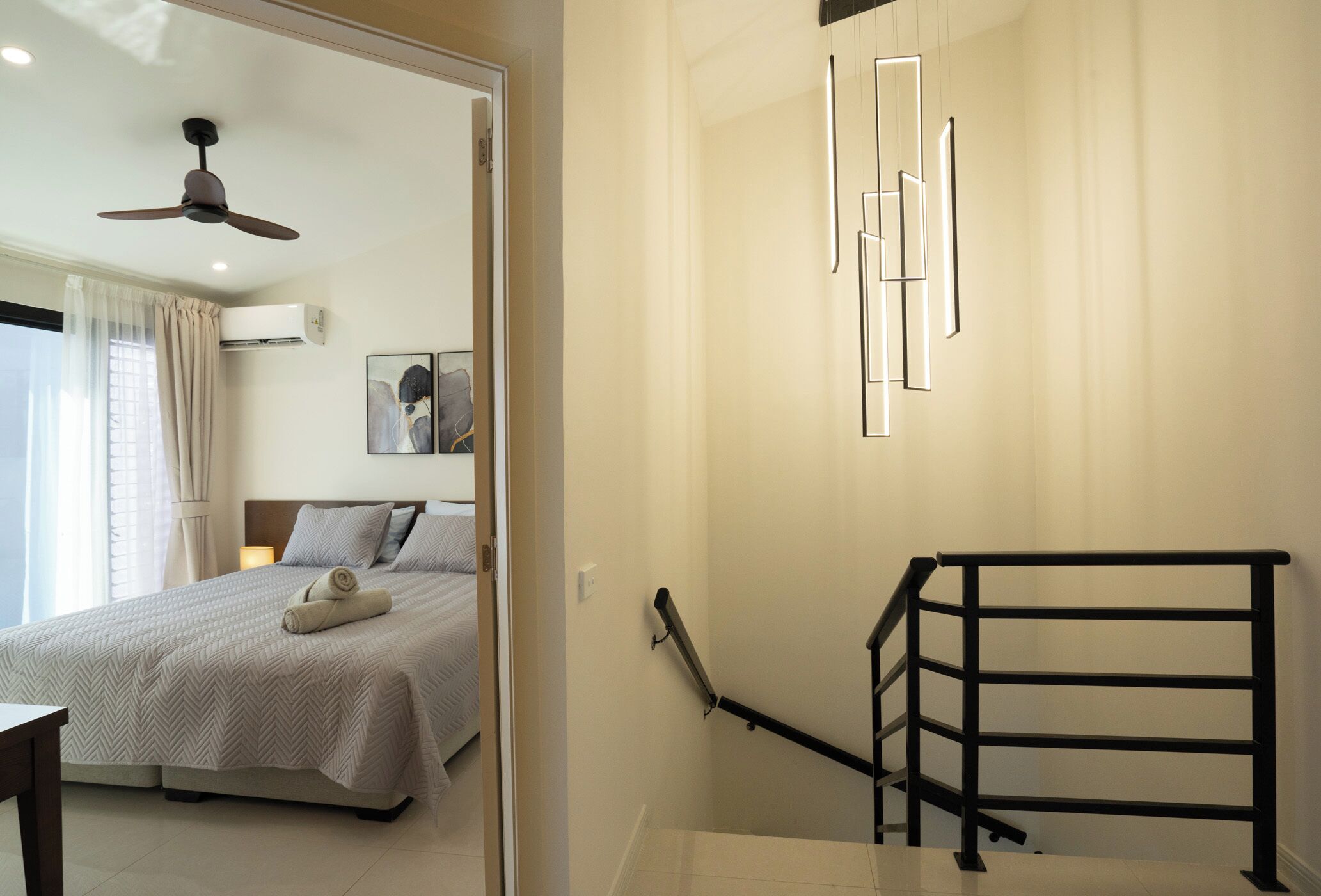 A hotel room with a bed, a ceiling fan, a wall-mounted air conditioner, a staircase with a handrail, and a framed picture on the wall.