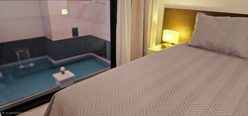 Quarto de hotel com cama, vista para a piscina e mesa de cabeceira com abajur.