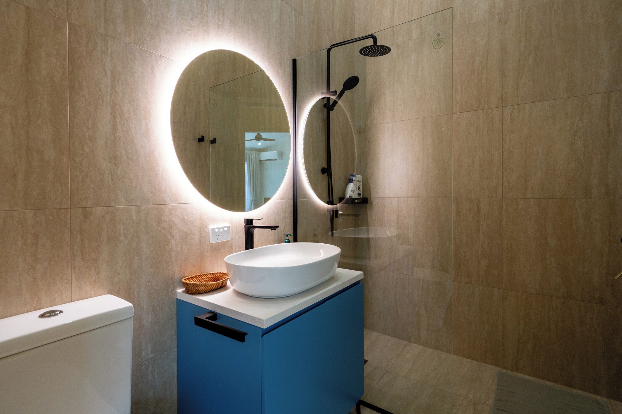 Un baño con un lavamanos blanco, un espejo con luz circular y una zona de ducha.