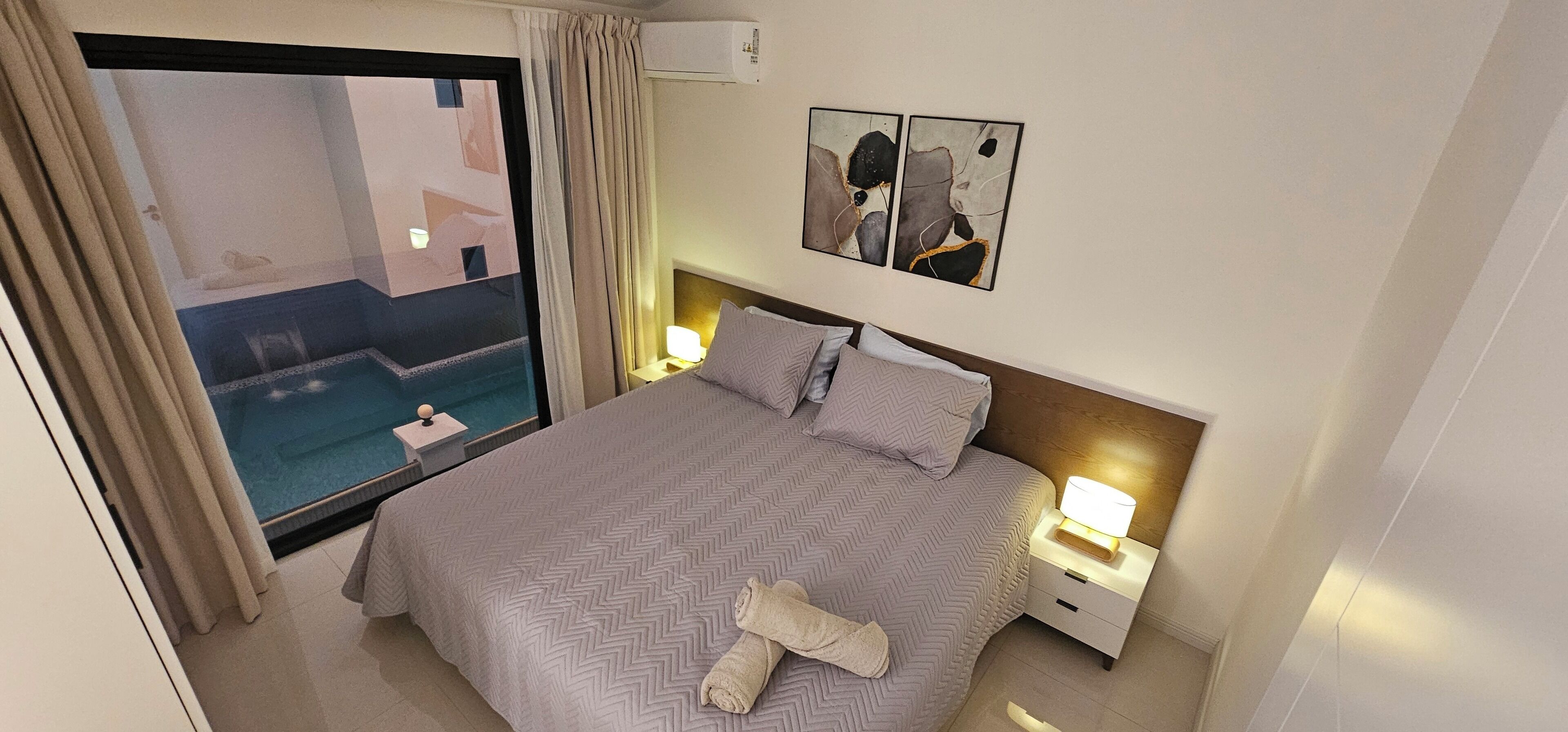 Une chambre d’hôtel comprenant un lit, une table de chevet, une lampe, une fenêtre avec des rideaux et un climatiseur fixé au mur.