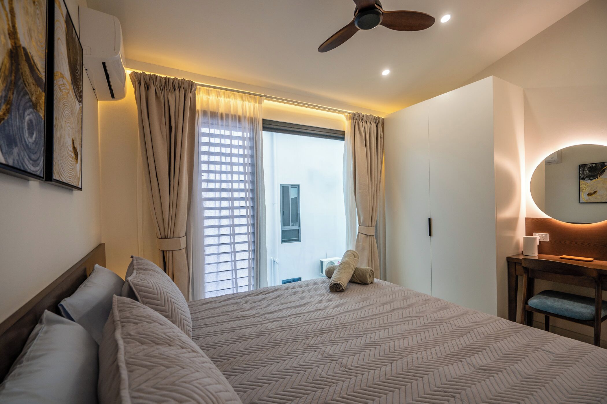 A hotel room with a bed, a ceiling fan, a desk with a chair, a window with curtains, and a wall-mounted air conditioner.