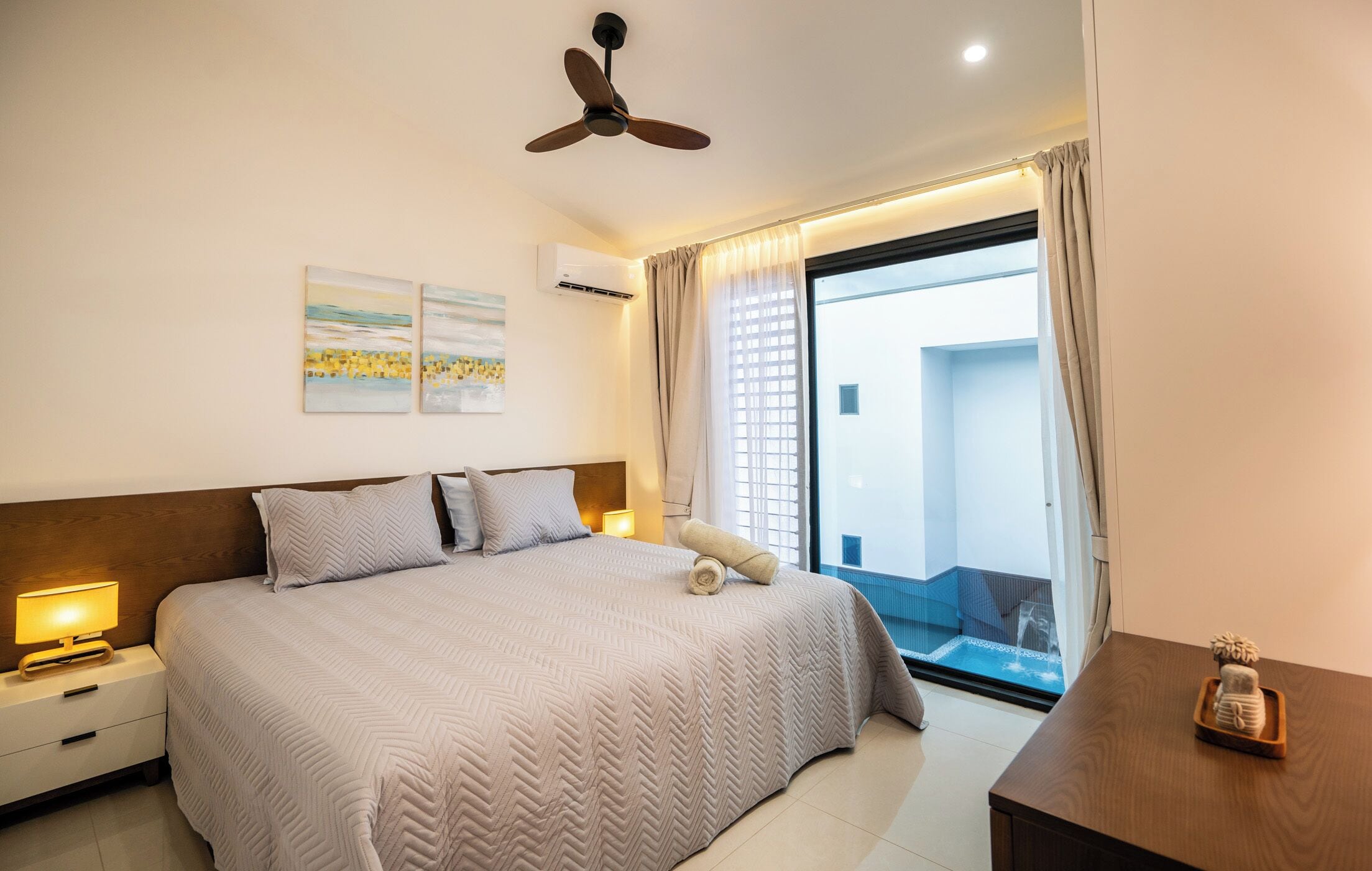 Habitación de hotel con una cama grande, ventilador de techo, mesita de noche con lámpara y vistas al balcón exterior.
