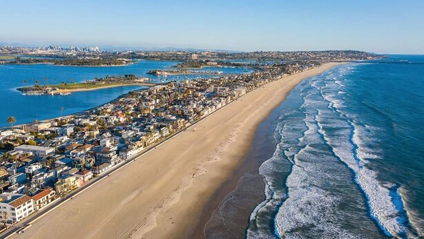 An aerial view of a coastal town with a long beachfront, residential buildings, and a marina. - Mission Beach Stay | A/C • Garage • Steps to Water (San Diego)