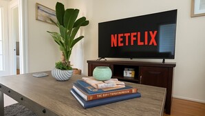 A living room with a wooden coffee table, a potted plant, a stack of books, and a television displaying the Netflix logo. - Mission Beach Stay | A/C • Garage • Steps to Water (San Diego)