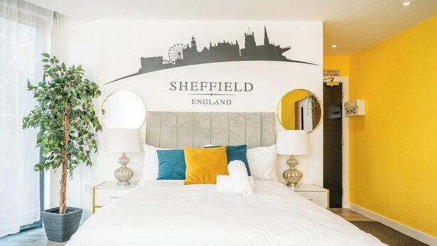 A hotel room with a bed, a city skyline mural, a potted plant, and a bright yellow wall. - Chic Studio | Prime Location | Free Parking (Sheffield)