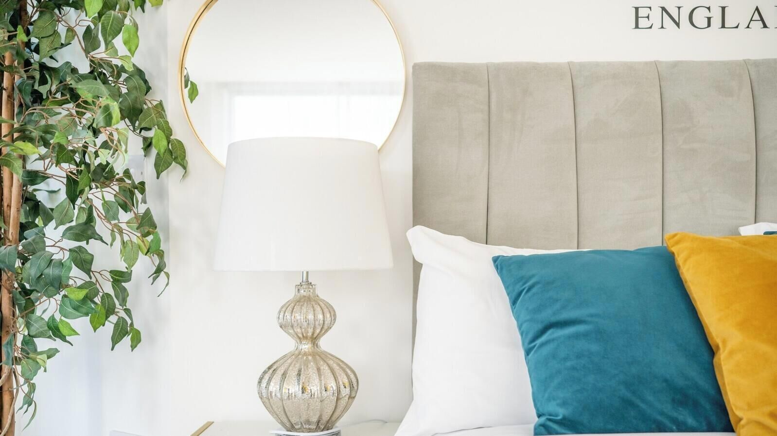 Un dormitorio ordenado con cama, lámpara de noche y un espejo instalado en la pared.