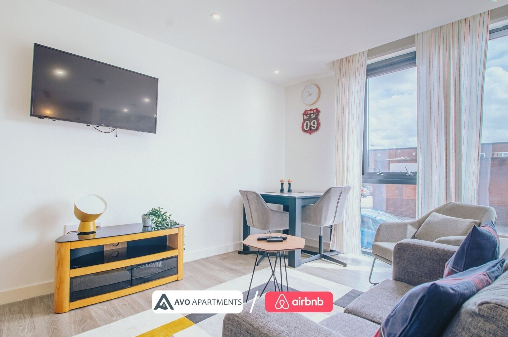 Een moderne woonkamer met een flatscreen televisie, een houten tv-meubel, een tafeltje met een plant en een bank. Er is een eethoek met een tafel en stoelen, en een groot raam met gordijnen.