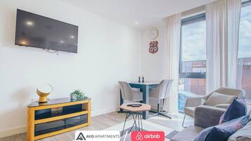 Een moderne woonkamer met een flatscreen televisie, een houten tv-meubel, een tafeltje met een plant en een bank. Er is een eethoek met een tafel en stoelen, en een groot raam met gordijnen.