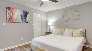 A bedroom with a bed, a ceiling fan, a door, a wall art piece, a lamp, and a decorative wall plate.