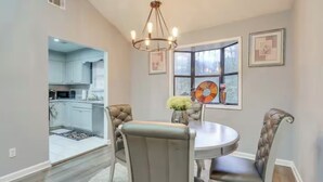 A dining area with a round table, cushioned chairs, a chandelier, and a window with a view of the kitchen.