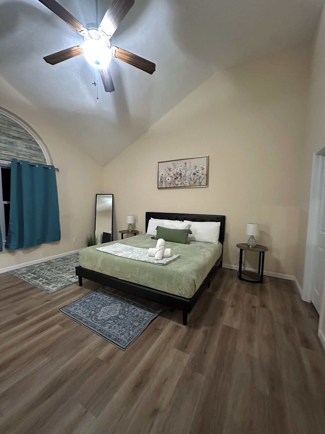 A bedroom with a bed, a ceiling fan, a painting, and a small table with a lamp.