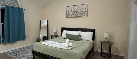 A bedroom with a bed, a ceiling fan, a painting, and a small table with a lamp.