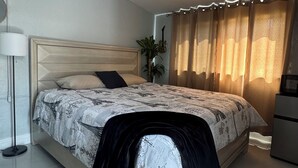 A bedroom with a bed, a bedside lamp, a dresser, and a window with curtains. - Private Chic Studio with Patio • King Bed • Near Beach (Hollywood)
