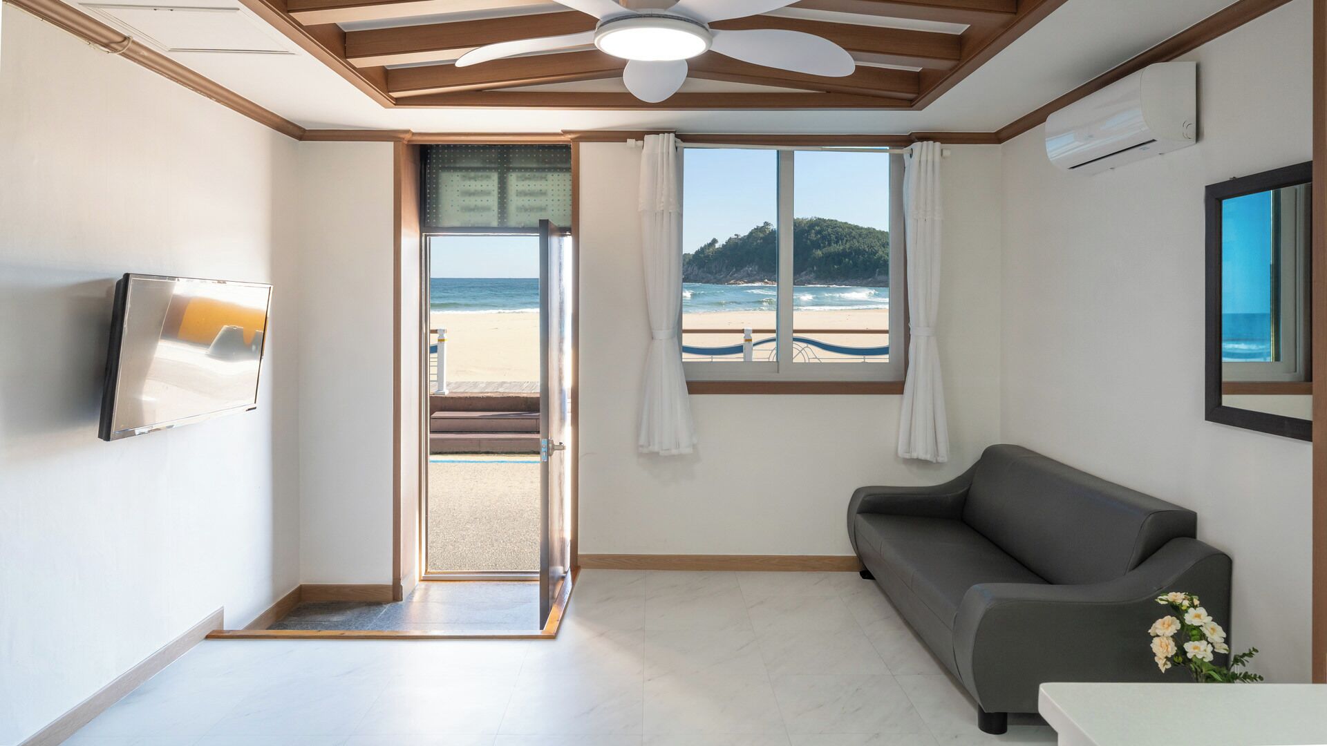 Habitación con vista al mar, ventilador de techo, televisor de pantalla plana y sofá gris.