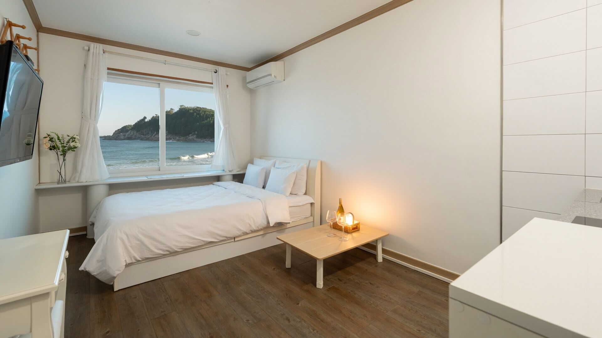 Un dormitorio con cama, mesita de noche con vasos, ventana con vista al mar y aire acondicionado.