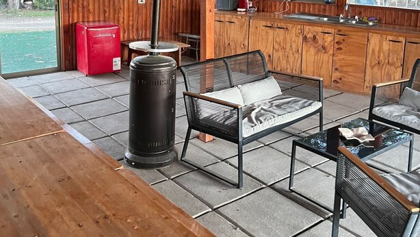A covered outdoor seating area with a wooden table, two black metal chairs, a glass coffee table, and a black heater. There is a red cooler and a window with a view of trees and a body of water. - CasaBosque minutes from the sea. (Placilla)