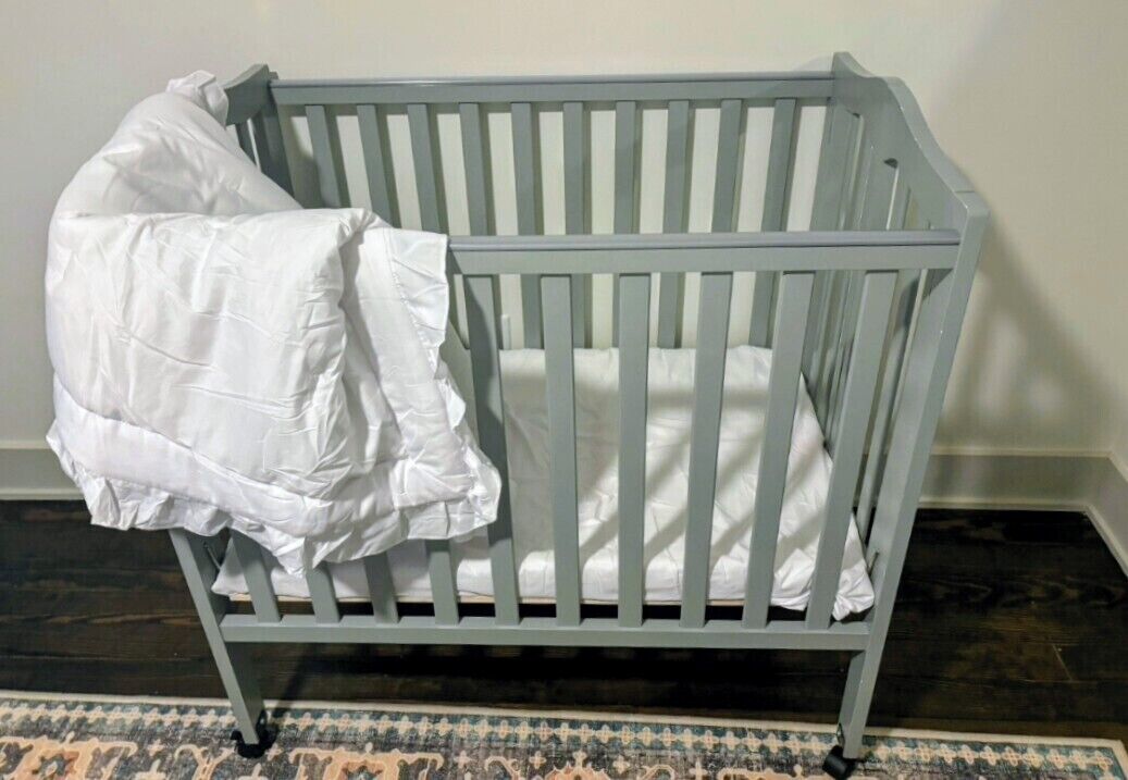 Una cuna gris con una manta blanca sobre ella, ubicada frente a una pared con un tapete estampado en el suelo.