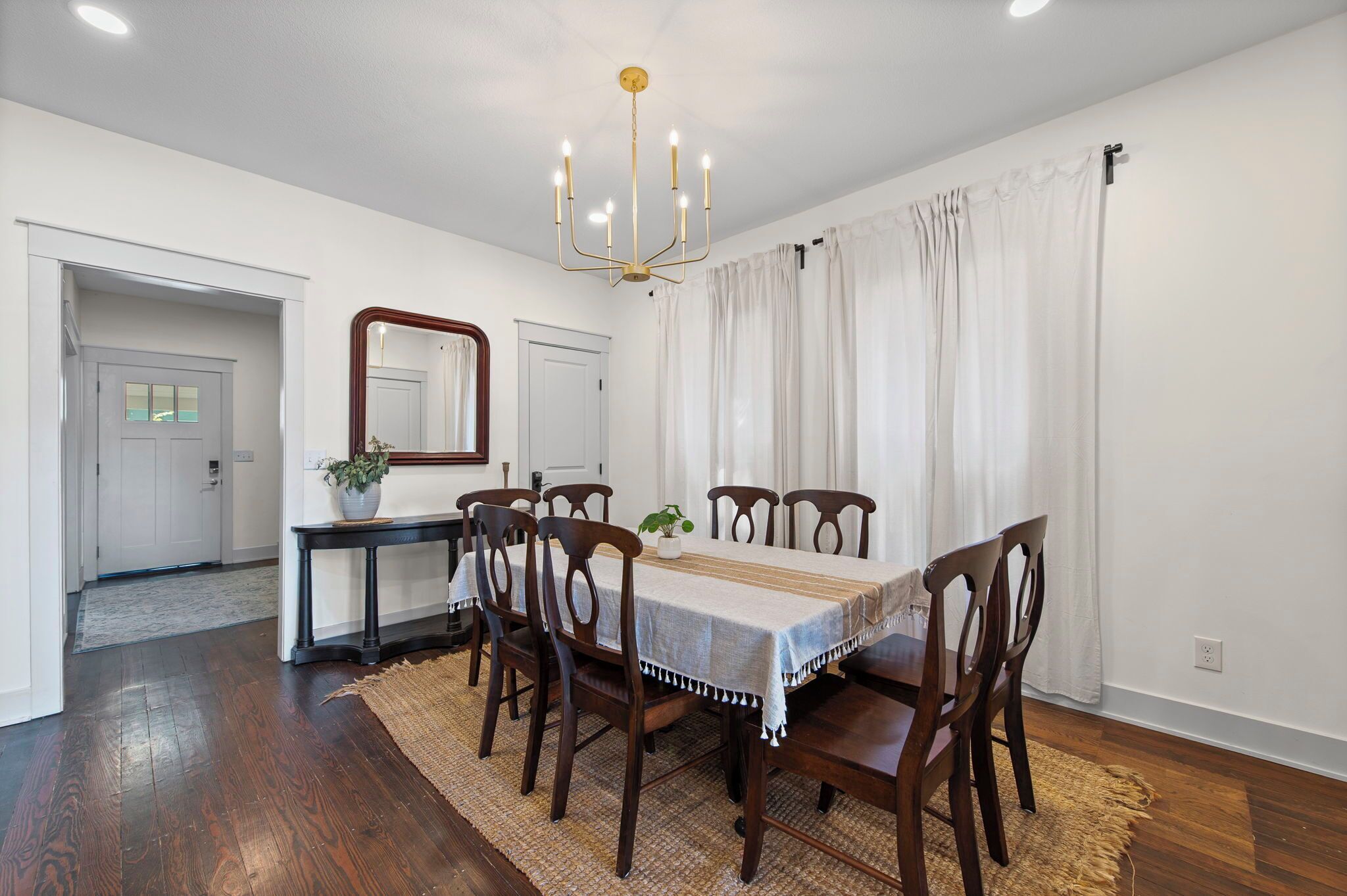 Comedor con mesa y sillas de madera, una araña y cortinas blancas.