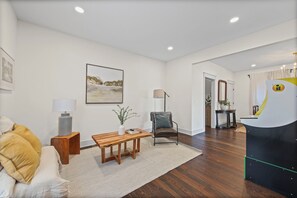 Ein modernes Wohnzimmer mit Couch, Sessel, Holztisch und Lampe. An der Wand hängt ein Bild und auf einem kleinen Tisch steht eine Pflanze.