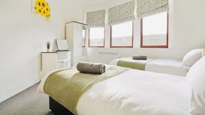 Quarto de hotel com duas camas, uma escrivaninha, um radiador e uma janela com cortinas.