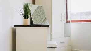 Uma pequena mesa com um laptop e uma planta em vaso, um armário branco e uma janela com vista.