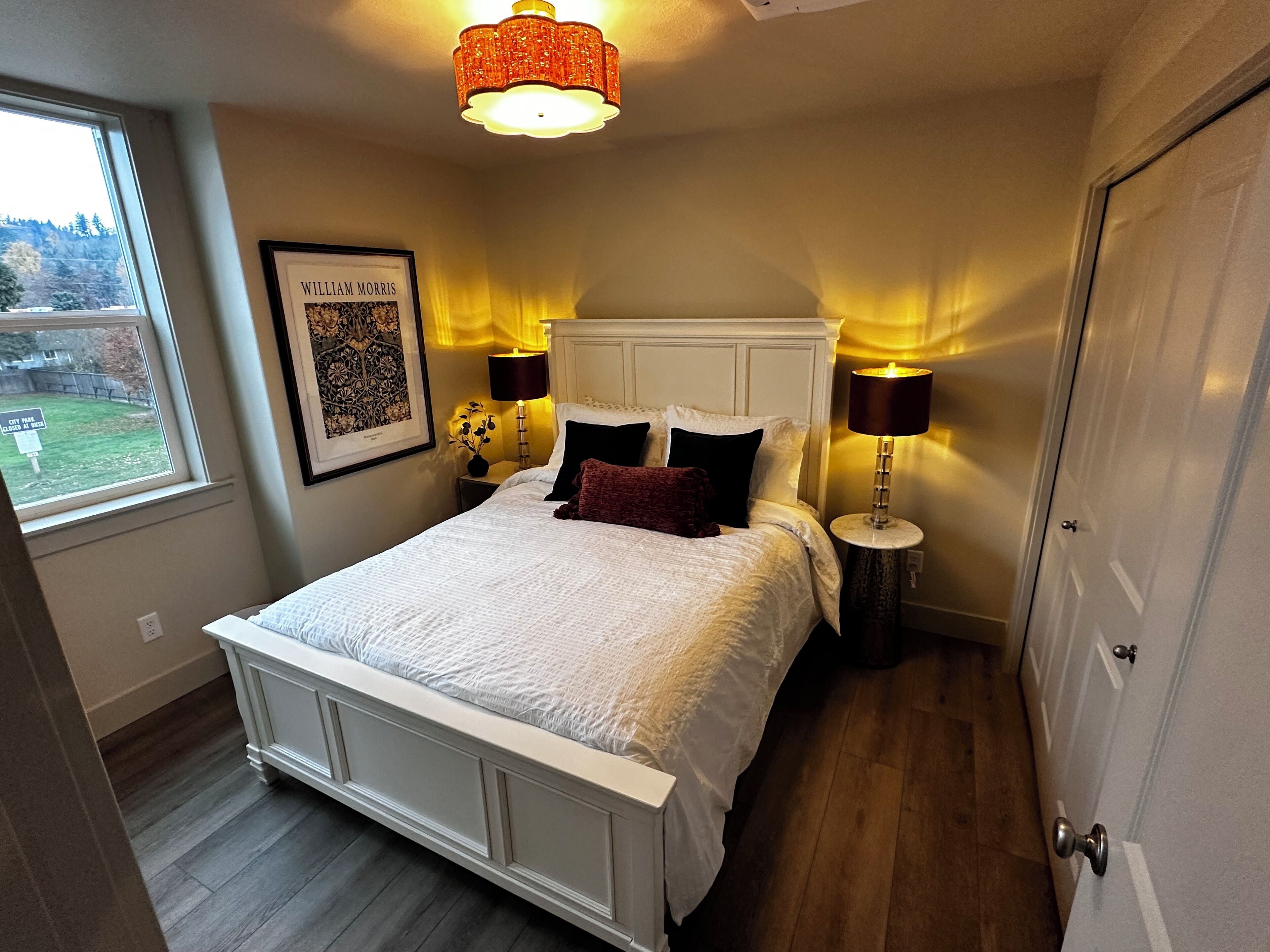 A bedroom with a white bed, two bedside tables with lamps, a framed picture on the wall, and a window with a view of trees.
