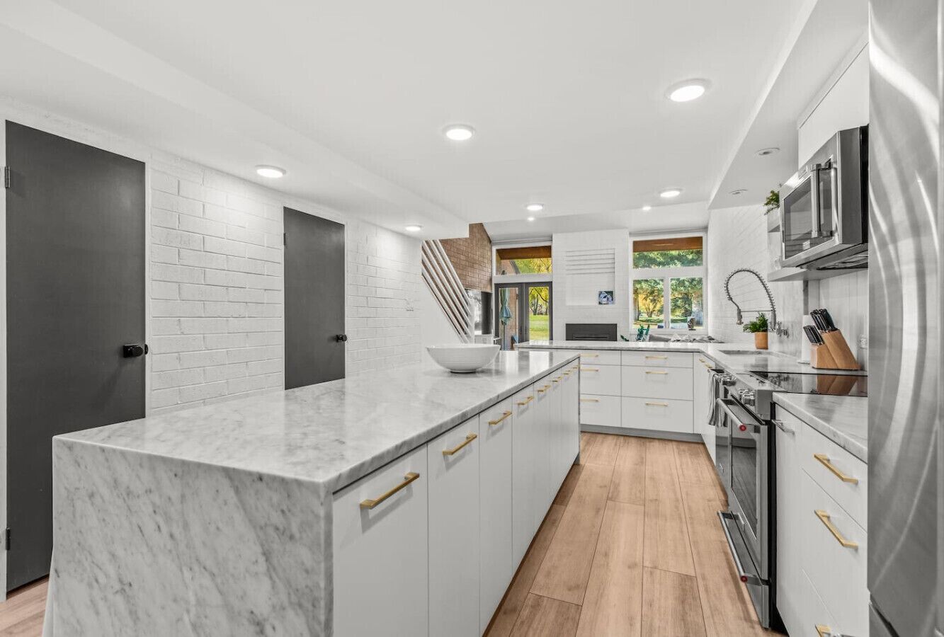 A modern kitchen with white cabinetry, a marble island, stainless steel appliances, and a wooden floor.