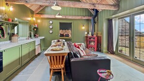 Ein gemütliches Interieur mit Holzbalkendecke, einem Essbereich mit langem Tisch und Stühlen, einer Couch und einem Kamin aus Stein.