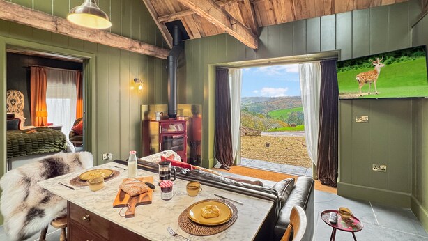 Ein gemütliches Zimmer mit Holzbalkendecke, Kamin, gedecktem Esstisch mit Speisen und einem Fernseher, der einen Hirsch zeigt.