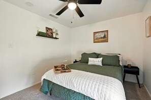 Un dormitorio con cama, mesita de noche, ventilador de techo, estante en la pared con objetos decorativos e imagen enmarcada.